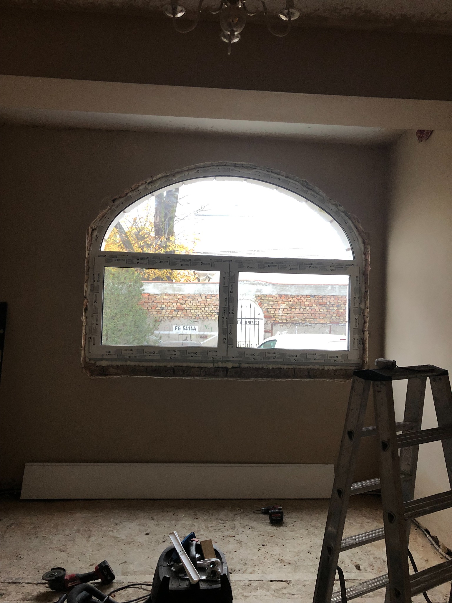 Montaż okna o nietypowym, łukowym kształcie w surowym wnętrzu. Widoczne narzędzia pracy i drabina. Na zewnątrz budynku widoczny fragment ceglanego muru i zaparkowany samochód.