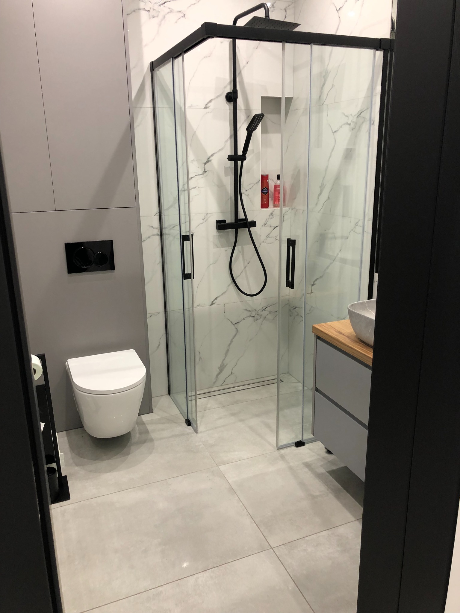 Nowoczesna łazienka z kabiną prysznicową typu walk-in z czarnymi profilami, podwieszaną toaletą i umywalką nablatową na drewnianym blacie.