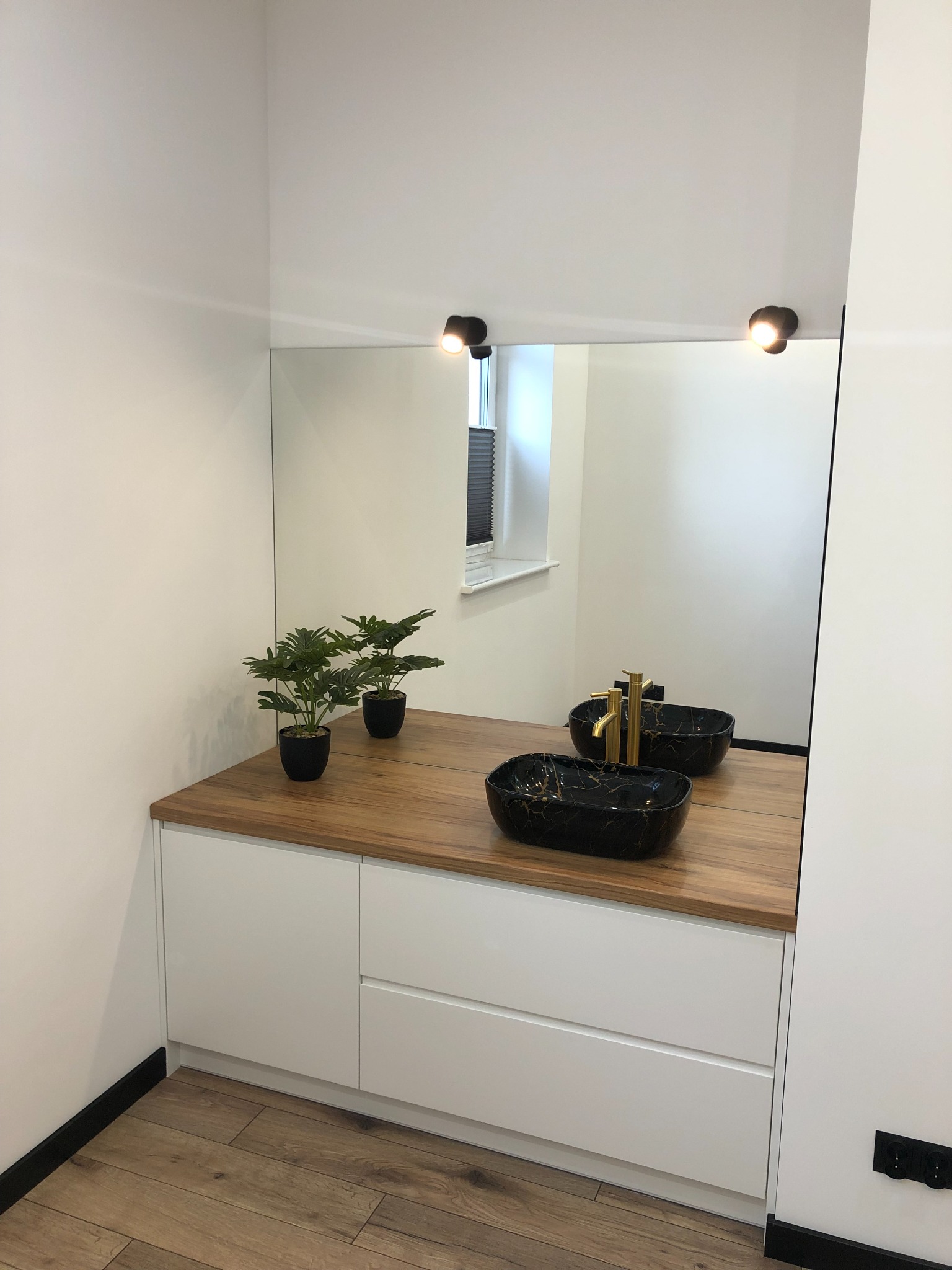 Nowoczesna łazienka z podwójną, czarną umywalką z marmurowym wzorem i złotą armaturą, blatem z drewna, białymi szafkami i lustrem z oświetleniem punktowym.