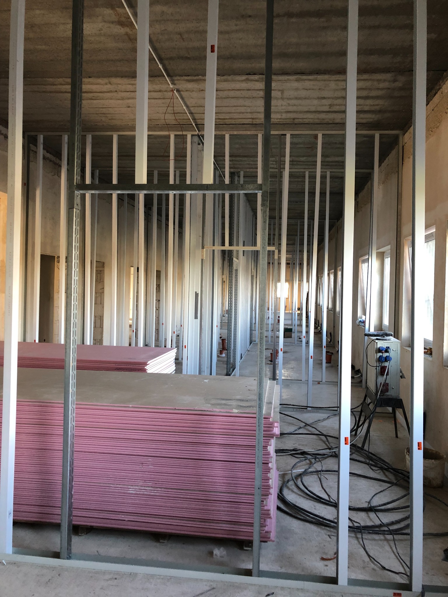 Stelaż z profili stalowych przygotowany pod montaż płyt gipsowo-kartonowych; na pierwszym planie stos różowych płyt GK, w tle widoczne okna i przewody elektryczne.