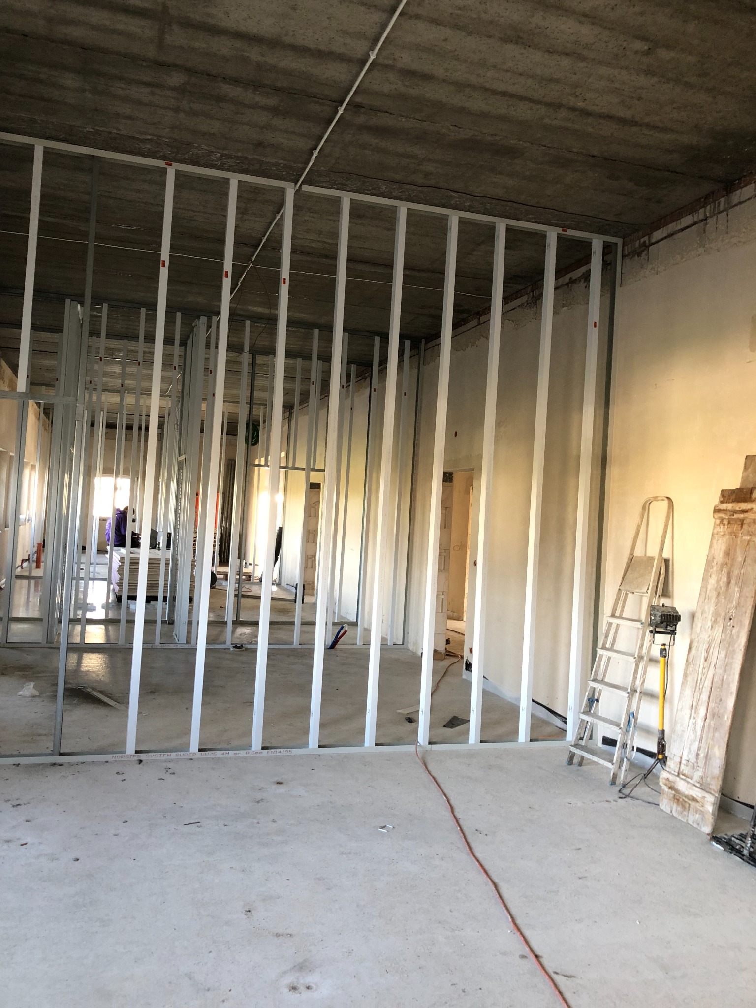 Montaż metalowego stelażu pod ścianę działową z płyty gipsowo-kartonowej w surowym wnętrzu, widoczna drabina i przewód elektryczny na podłodze.