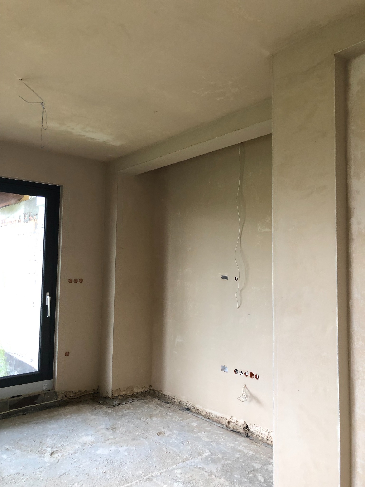 Wnętrze w trakcie prac wykończeniowych: ściany pokryte świeżym tynkiem, widoczne gniazdka elektryczne i wystające przewody, surowa podłoga.
