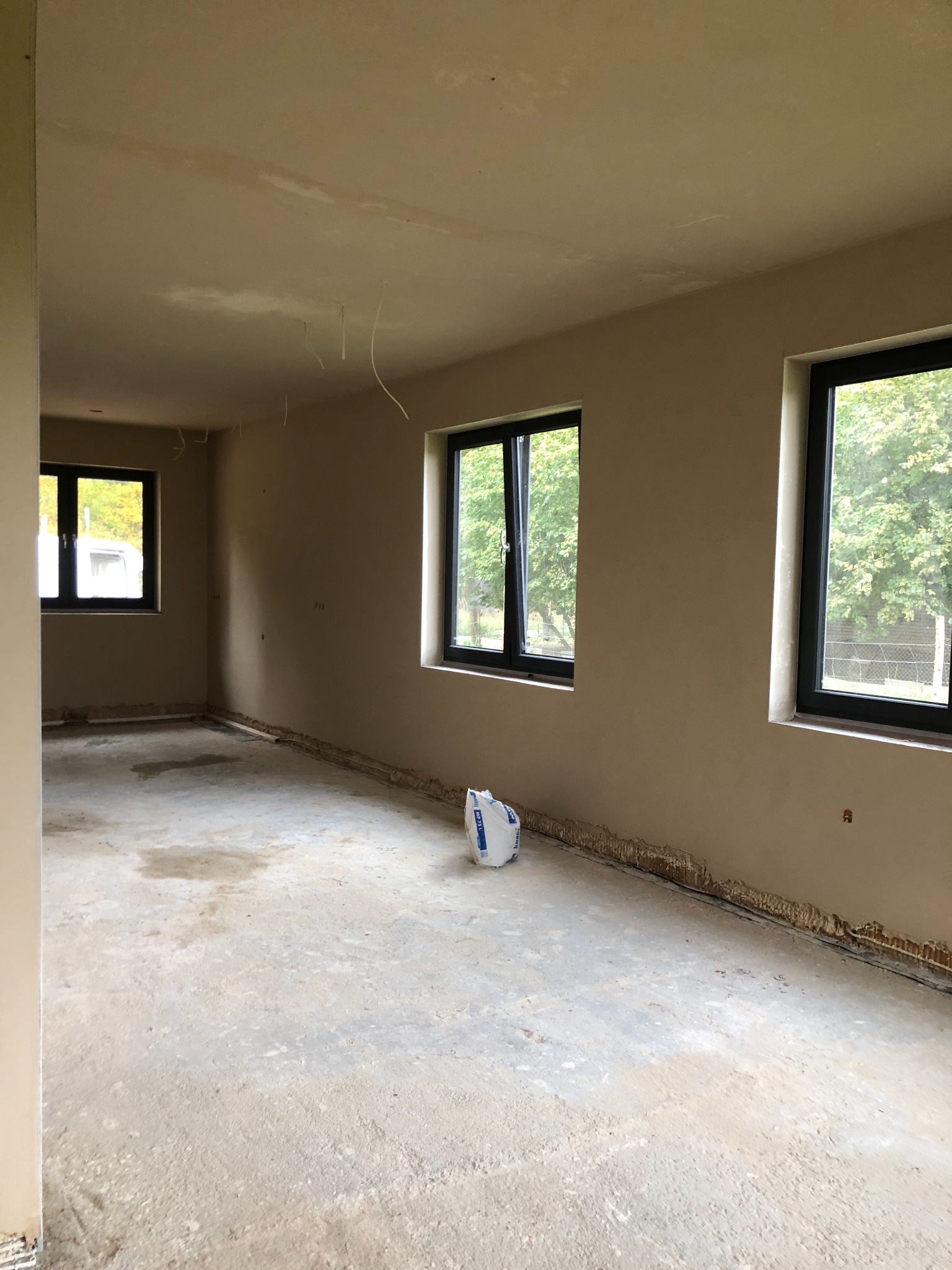 Surowe wnętrze w trakcie prac wykończeniowych: gołe ściany pokryte gładzią, betonowa podłoga, wystające przewody elektryczne z sufitu i worek gipsu przy ścianie z czarnymi oknami.