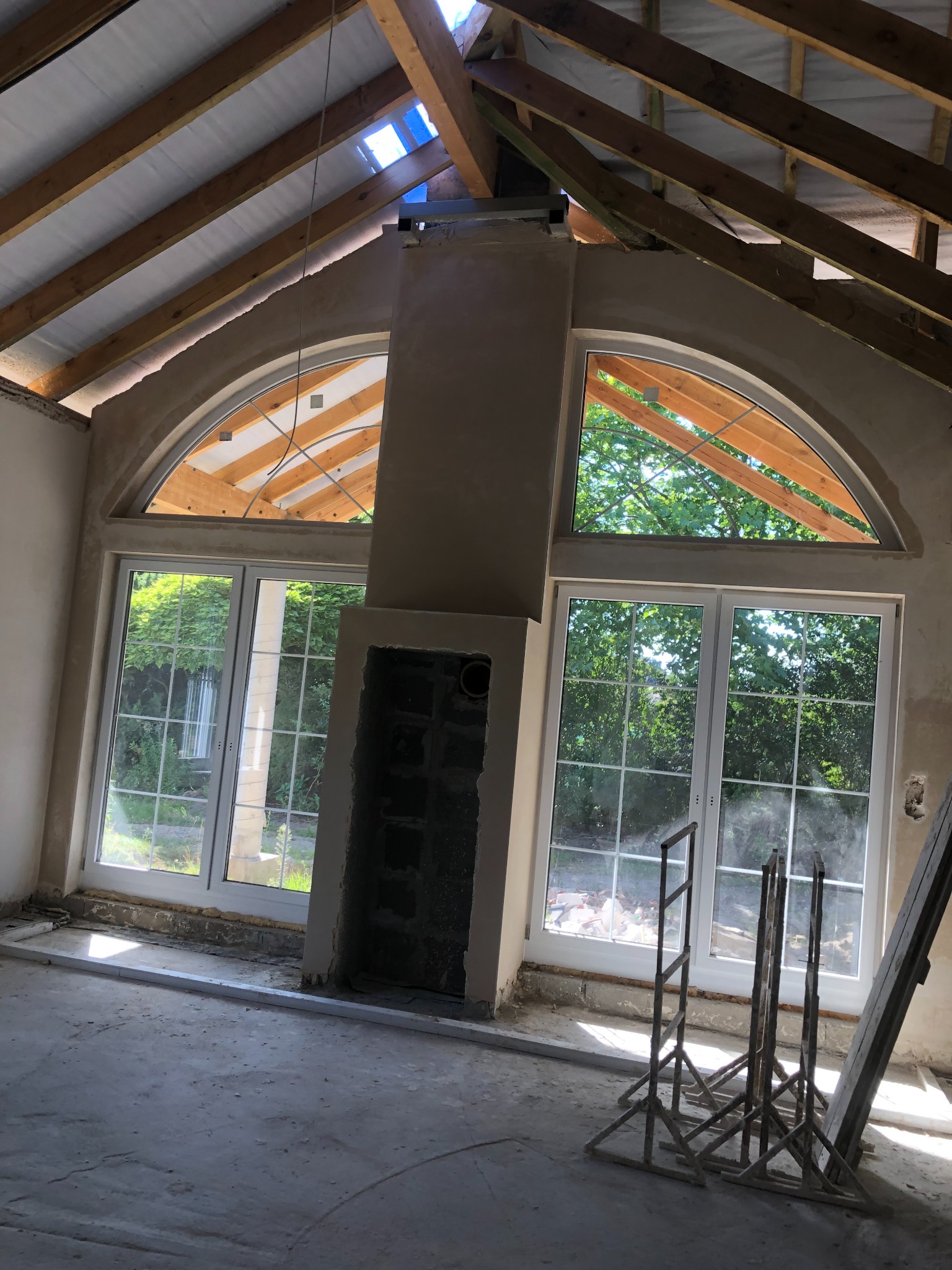 Wnętrze w trakcie prac wykończeniowych: surowy komin, duże okna łukowe z widokiem na zieleń, widoczne elementy konstrukcji dachu z drewnianych belek, podłoga w stanie surowym, widoczne podpory...