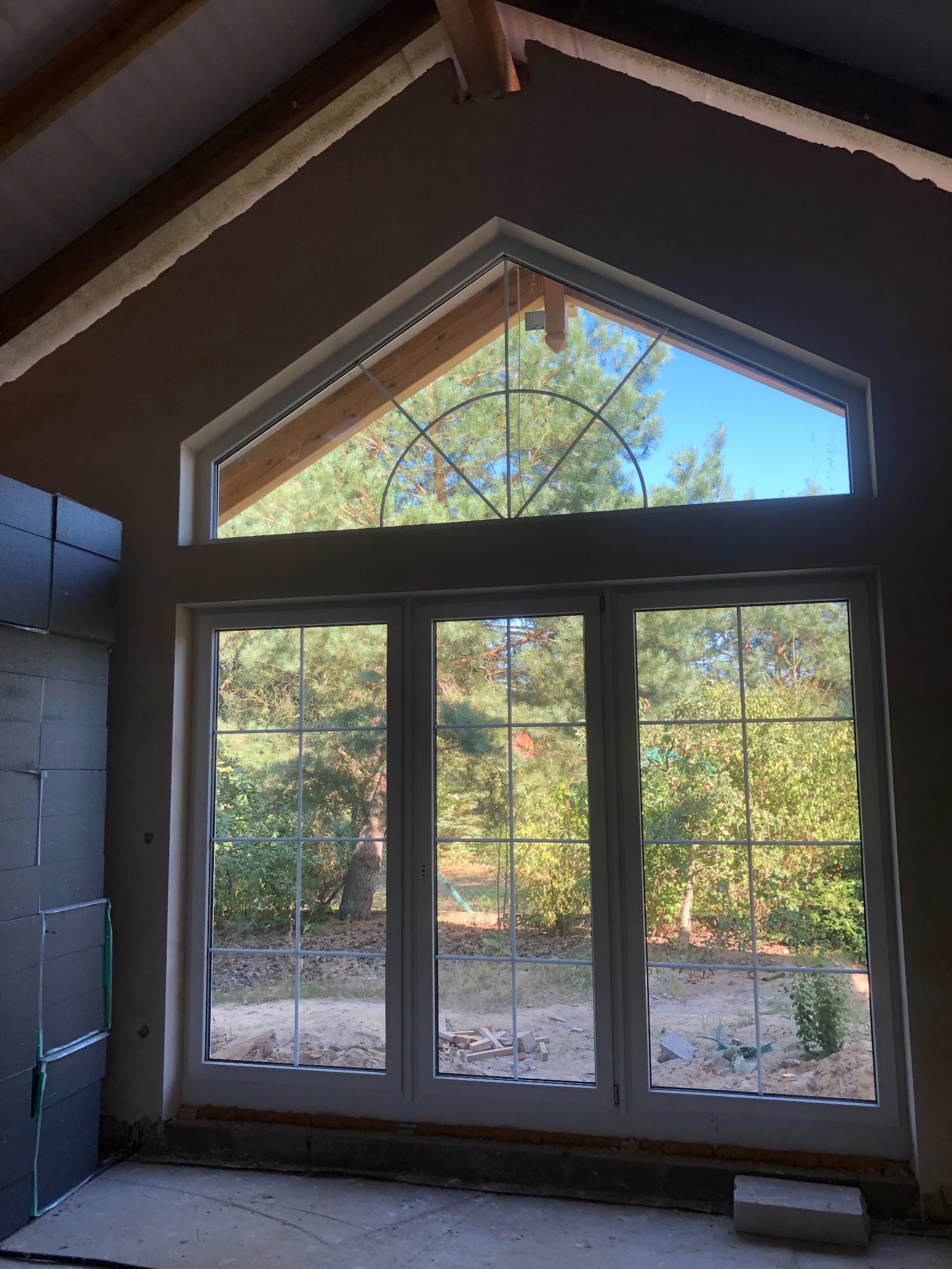 Nowe, białe okno trzyskrzydłowe z ozdobnym, półokrągłym nadświetlem w kształcie wachlarza, zamontowane w surowym wnętrzu w trakcie prac wykończeniowych.