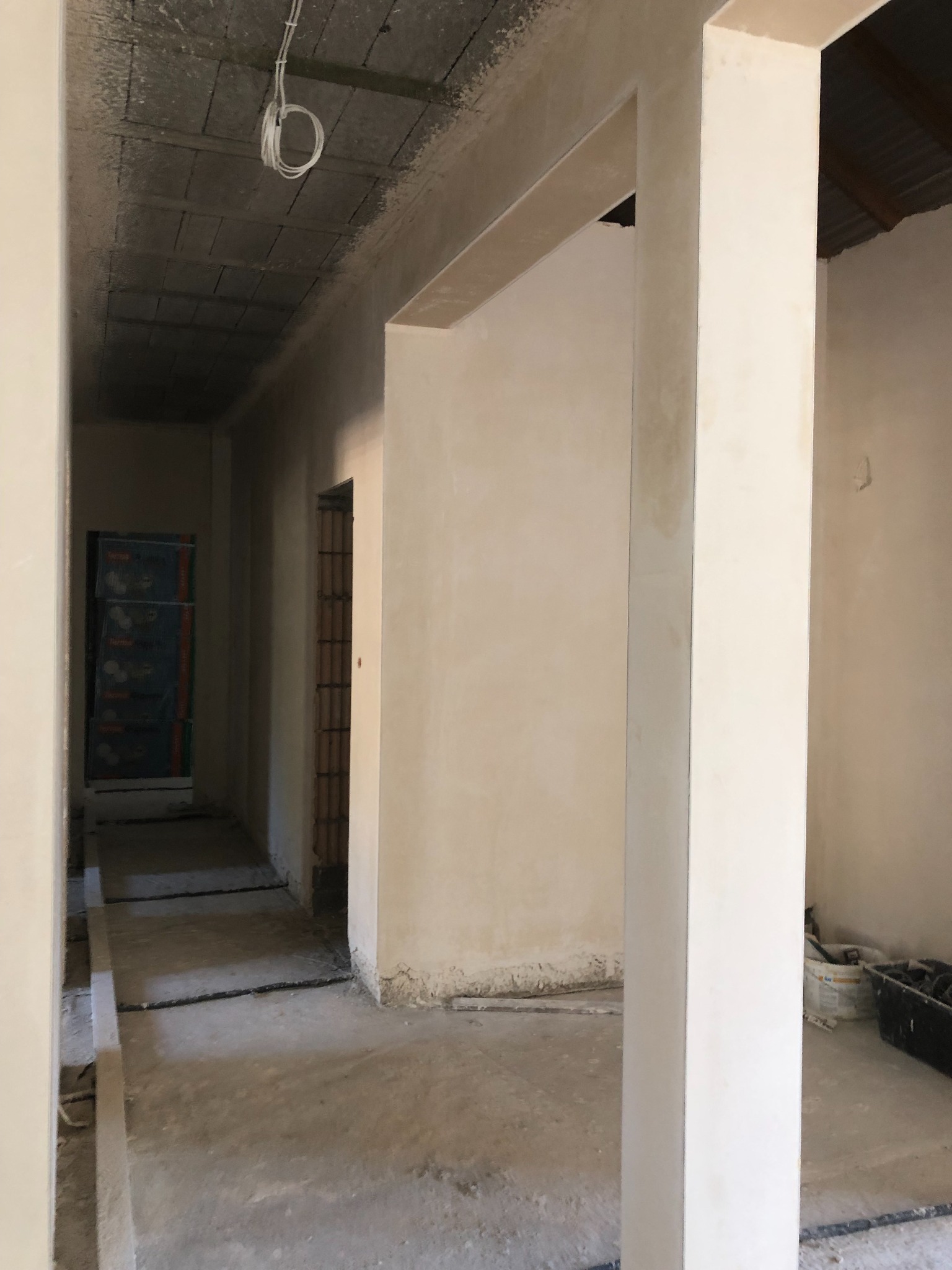 Korytarz w trakcie prac wykończeniowych, widoczne świeżo otynkowane ściany, surowy betonowy strop z wystającymi kablami i podłoga w trakcie przygotowania.