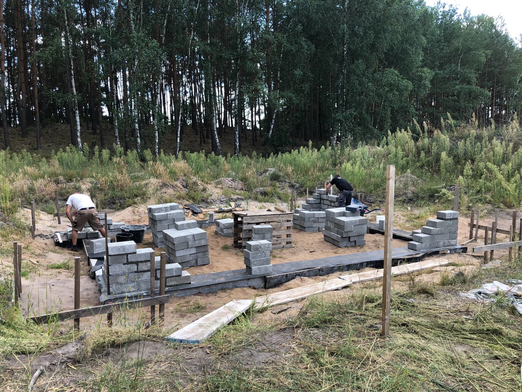 Budowa fundamentów domu z bloczków betonowych na piaszczystym terenie, widoczni dwaj pracownicy w trakcie murowania, w tle las.