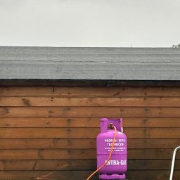 METAL-TRANS Mariusz Błoński - Różowa butla z gazem propan-butan na tle drewnianej ściany budynku z dachem pokrytym papą.