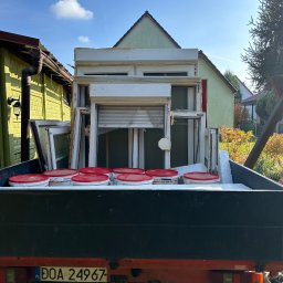 METAL-TRANS Mariusz Błoński - Ciężarówka z otwartą skrzynią ładunkową wypełnioną starymi oknami, roletami i wiadrami z farbą z czerwonymi pokrywkami, na tle domów w słoneczny dzień.