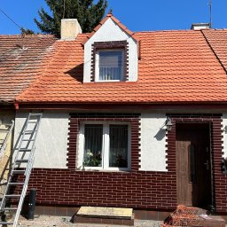 METAL-TRANS Mariusz Błoński - Dom z częściowo wymienionym pokryciem dachu z dachówki ceramicznej, widoczna drabina oparta o ścianę i narzędzia na chodniku przed domem.