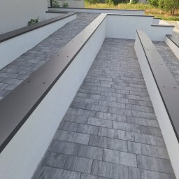 METAL-TRANS Mariusz Błoński - Nowoczesny podjazd z szarej kostki brukowej, ograniczony niskimi, białymi murkami z metalowymi listwami, prowadzący do budynku z jasną elewacją.