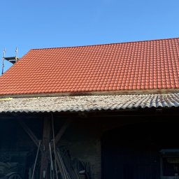 METAL-TRANS Mariusz Błoński - Czerwony dach z dachówki ceramicznej po wymianie, widoczny fragment starego dachu z eternitu oraz rusztowanie przy kominie na tle błękitnego nieba.