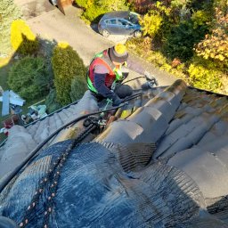 METAL-TRANS Mariusz Błoński - Pracownik w kasku i kamizelce, zabezpieczony linami, maluje dachówkę na ciemny kolor za pomocą pistoletu natryskowego. Widok z góry na częściowo pomalowany dach.