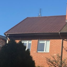 METAL-TRANS Mariusz Błoński - Ciemnobrązowy dach z blachy trapezowej na ceglanym domu z kominem i anteną, widok z dołu w słoneczny dzień.