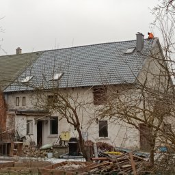 METAL-TRANS Mariusz Błoński - Pracownik w pomarańczowej kurtce na szarym dachu domu z lukarnami. W tle pochmurne niebo, a na pierwszym planie gałęzie drzew i resztki śniegu.