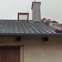 METAL-TRANS Mariusz Błoński - Dach z blachodachówki z oknem dachowym i kominem. Na dachu drabina dekarska. Widoczny fragment ściany budynku z drewnianymi drzwiami.