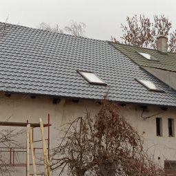 METAL-TRANS Mariusz Błoński - Szary dach z oknami dachowymi na białym budynku, widoczne rusztowania i drzewo bez liści. Część dachu ma inny, zielonkawy odcień.