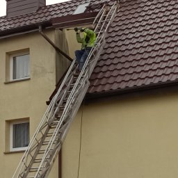 METAL-TRANS Mariusz Błoński - Pracownik w zielonej kurtce na drabinie czyści dach domu z brązową dachówką za pomocą myjki ciśnieniowej. Widoczne zabrudzenia na elewacji.