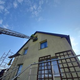 METAL-TRANS Mariusz Błoński - Pracownik na drabinie przy dachu domu z żółtą elewacją. Widoczne rusztowanie i błękitne niebo. Ujęcie z dołu, podkreślające wysokość budynku.