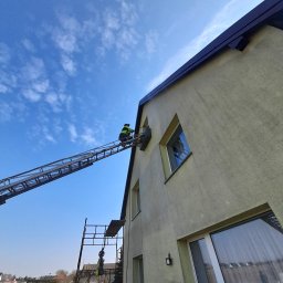 METAL-TRANS Mariusz Błoński - Pracownik na drabinie przy elewacji domu, prawdopodobnie montaż klimatyzacji. Widoczne rusztowanie i błękitne niebo.