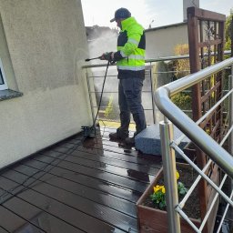 METAL-TRANS Mariusz Błoński - Mężczyzna w odblaskowej kurtce czyści elewację domu myjką ciśnieniową z balkonu. Widoczne zacieki i czysta powierzchnia. W tle fragment dachu i niebo.