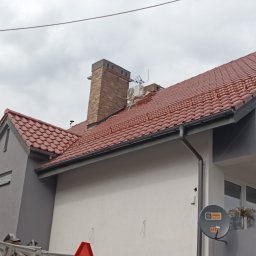 METAL-TRANS Mariusz Błoński - Czerwona dachówka na dachu domu z ceglanym kominem. Widoczna drabina z pracownikiem. Antena satelitarna na ścianie. Pochmurne niebo w tle.