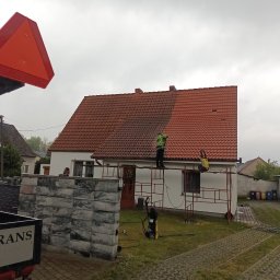 METAL-TRANS Mariusz Błoński - Mycie dachu z czerwonej dachówki. Część dachu umyta, widoczna różnica w kolorze. Pracownik na rusztowaniu używa myjki ciśnieniowej. Widok z dołu, pochmurne niebo.