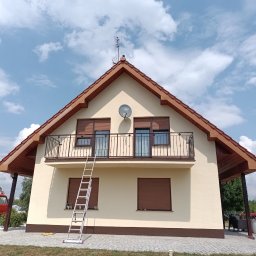 METAL-TRANS Mariusz Błoński - Beżowy dom z brązowym dachem dwuspadowym, anteną i talerzem satelitarnym na szczycie. Do elewacji oparta jest długa, aluminiowa drabina, a na balkonie widać metalową balustradę.