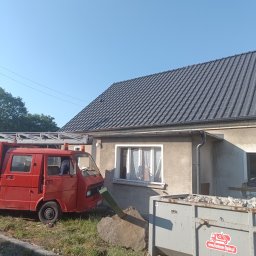 METAL-TRANS Mariusz Błoński - Nowy, ciemnoszary dach z dachówek i dopasowane rynny na beżowym domu jednorodzinnym, obok czerwona ciężarówka z drabiną i kontener na gruz z logo firmy Kontener-Opole.pl.