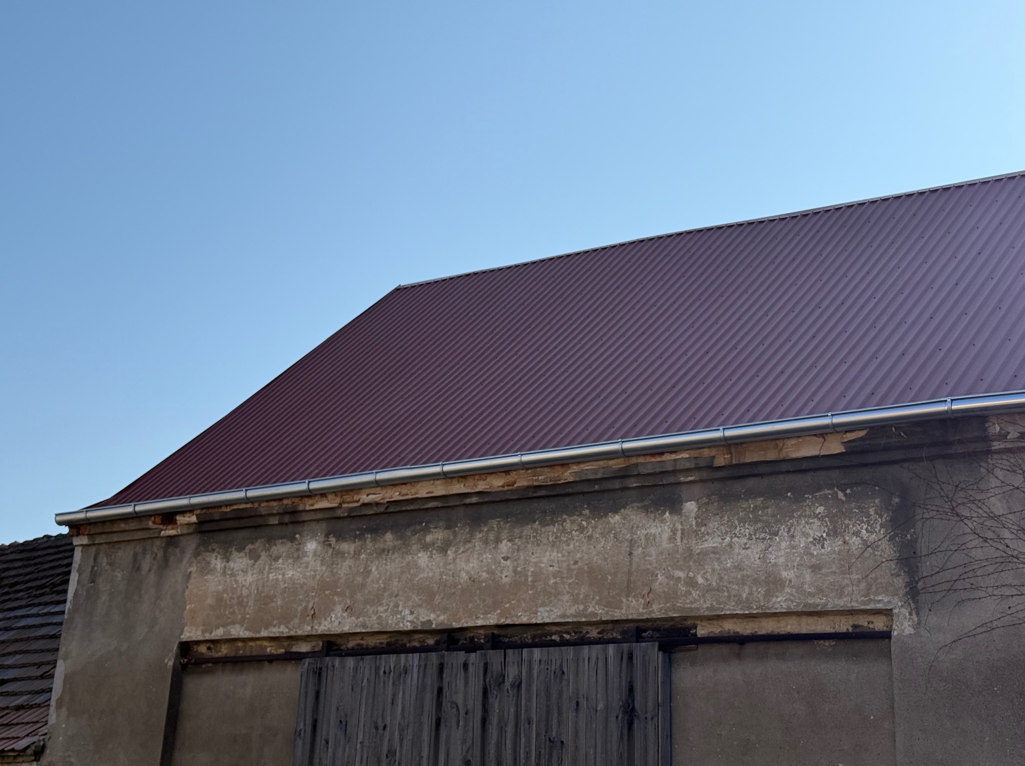 Czerwony, blaszany dach z zamontowaną rynną na szczycie starego, częściowo otynkowanego budynku z drewnianymi wrotami.