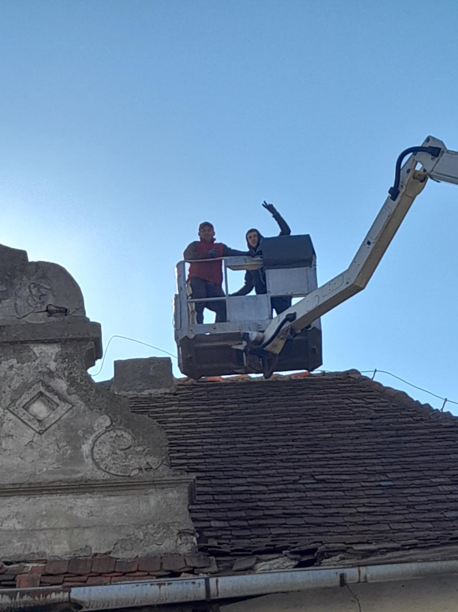 Dwóch pracowników na podnośniku koszowym na tle błękitnego nieba, pracujących na starym dachu z dachówki ceramicznej, widoczne detale architektoniczne na budynku.