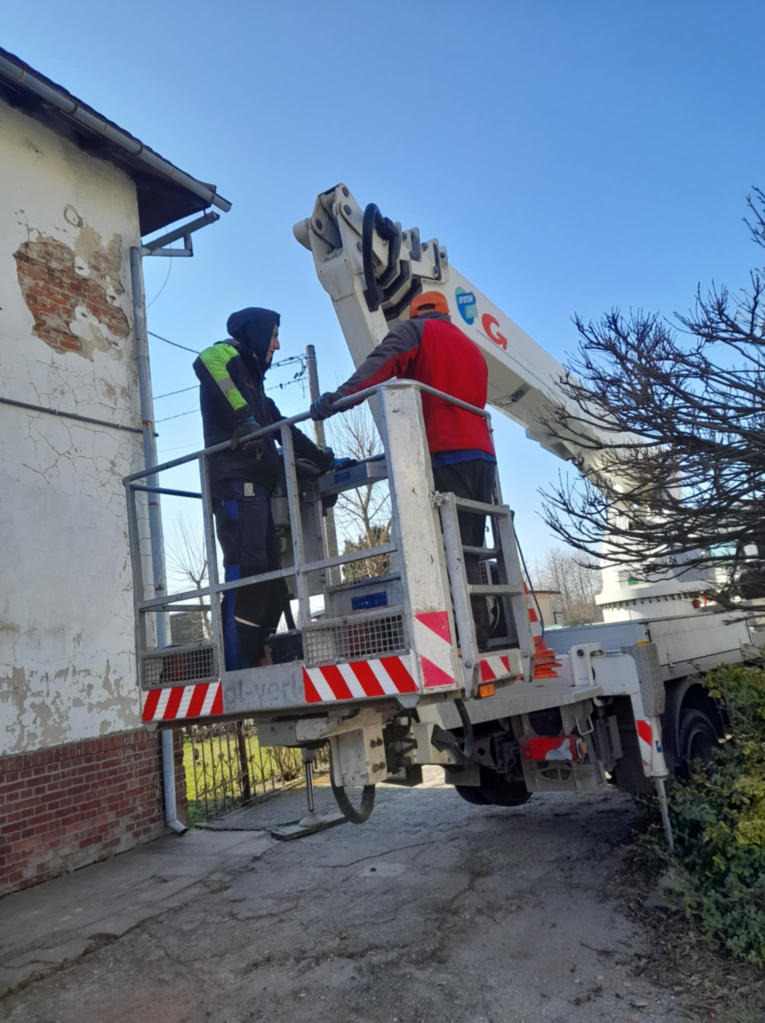 Dwóch pracowników na podnośniku koszowym przy budynku z uszkodzonym tynkiem, prawdopodobnie podczas prac związanych z elewacją lub dachem.