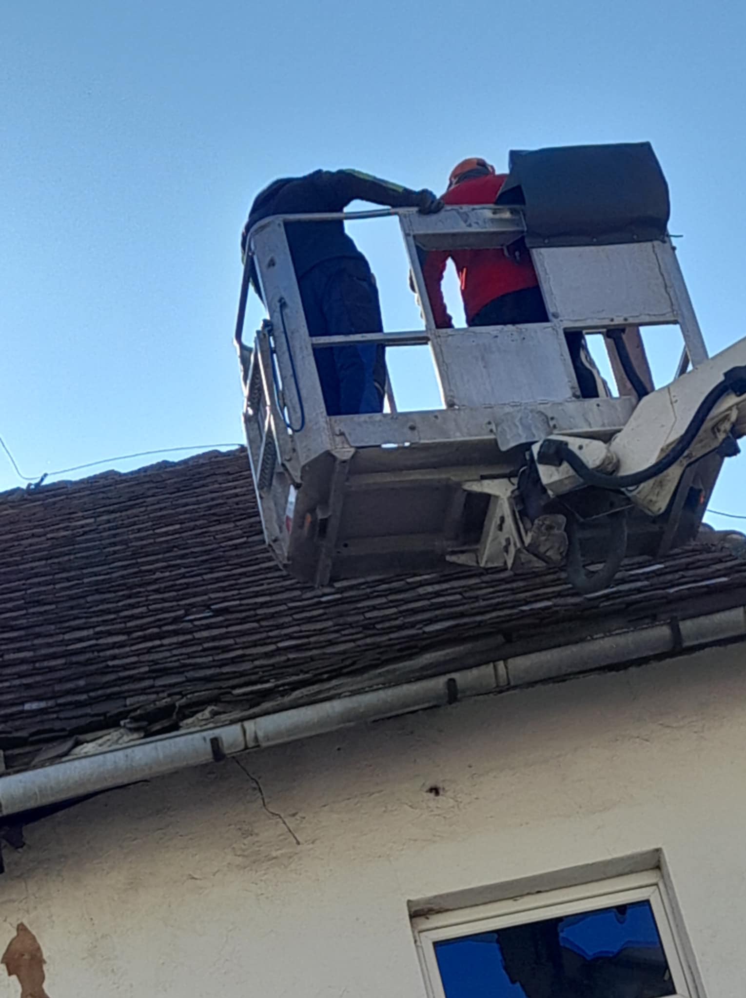 Dwóch pracowników w koszu podnośnika hydraulicznego, pracujących na starym, brązowym dachu z widocznymi uszkodzeniami, na tle błękitnego nieba.