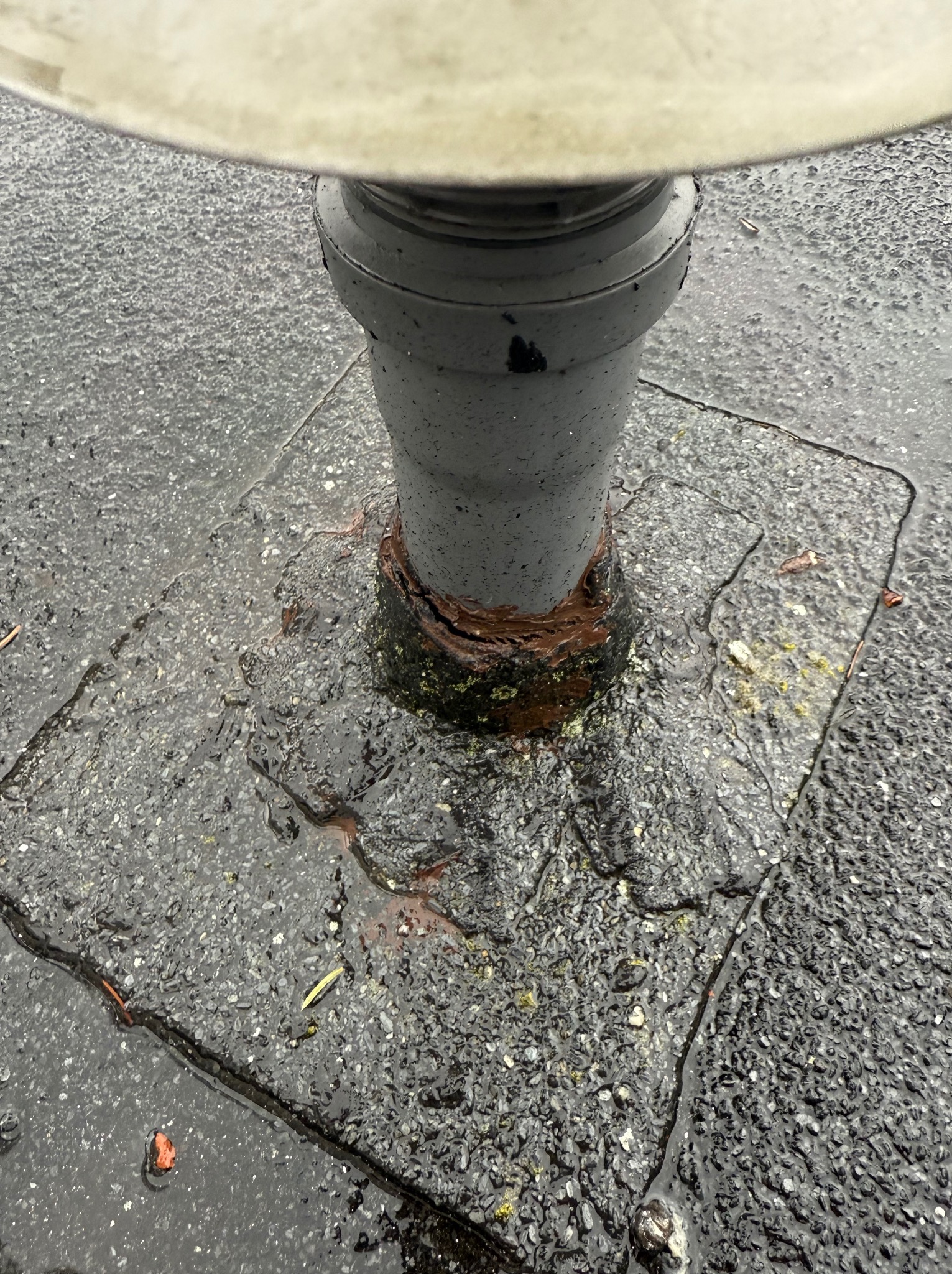 Szczegółowe ujęcie podstawy masztu oświetleniowego na mokrej, popękanej powierzchni z elementami mchu i brudu, z widocznymi śladami korozji i uszkodzeń. Woda zbiera się w zagłębieniach.