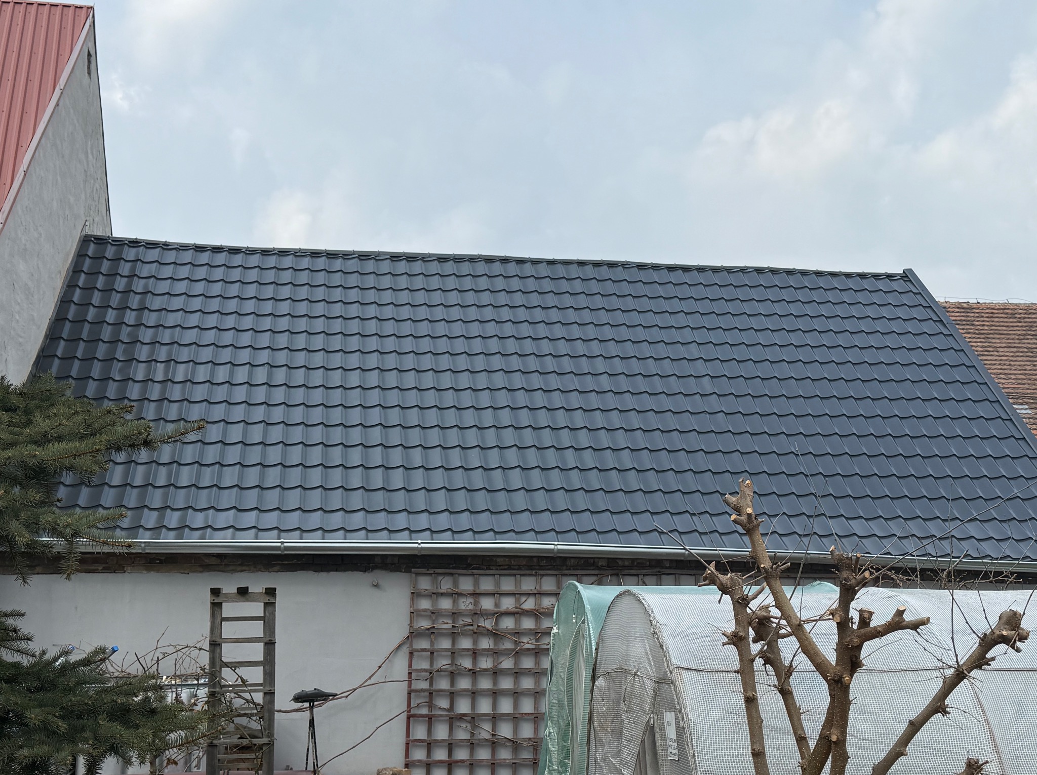 Widok na nowy, grafitowy dach z blachodachówki, z widoczną drabinką opartą o ścianę budynku i fragmentami ogrodu w tle.