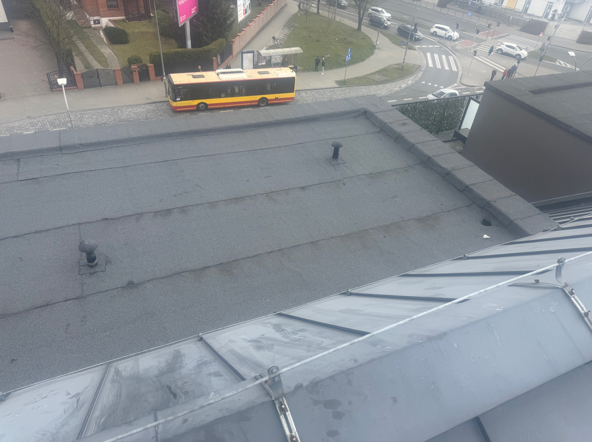 Widok z góry na płaski, szary dach pokryty papą, z widocznymi kominkami wentylacyjnymi i elementami obróbki blacharskiej, w tle ulica z autobusem i przejściem dla pieszych.