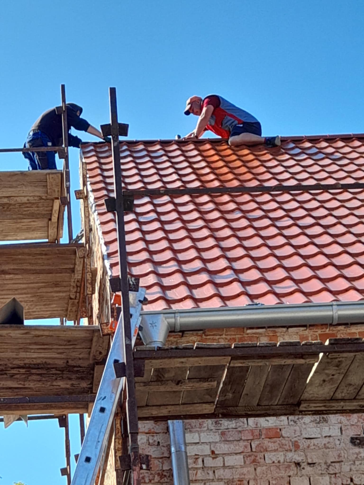 Dwóch dekarzy pracuje na dachu z czerwonej dachówki, używając rusztowania i drabiny. Widoczne rynny i ceglana ściana budynku. Błękitne niebo w tle.