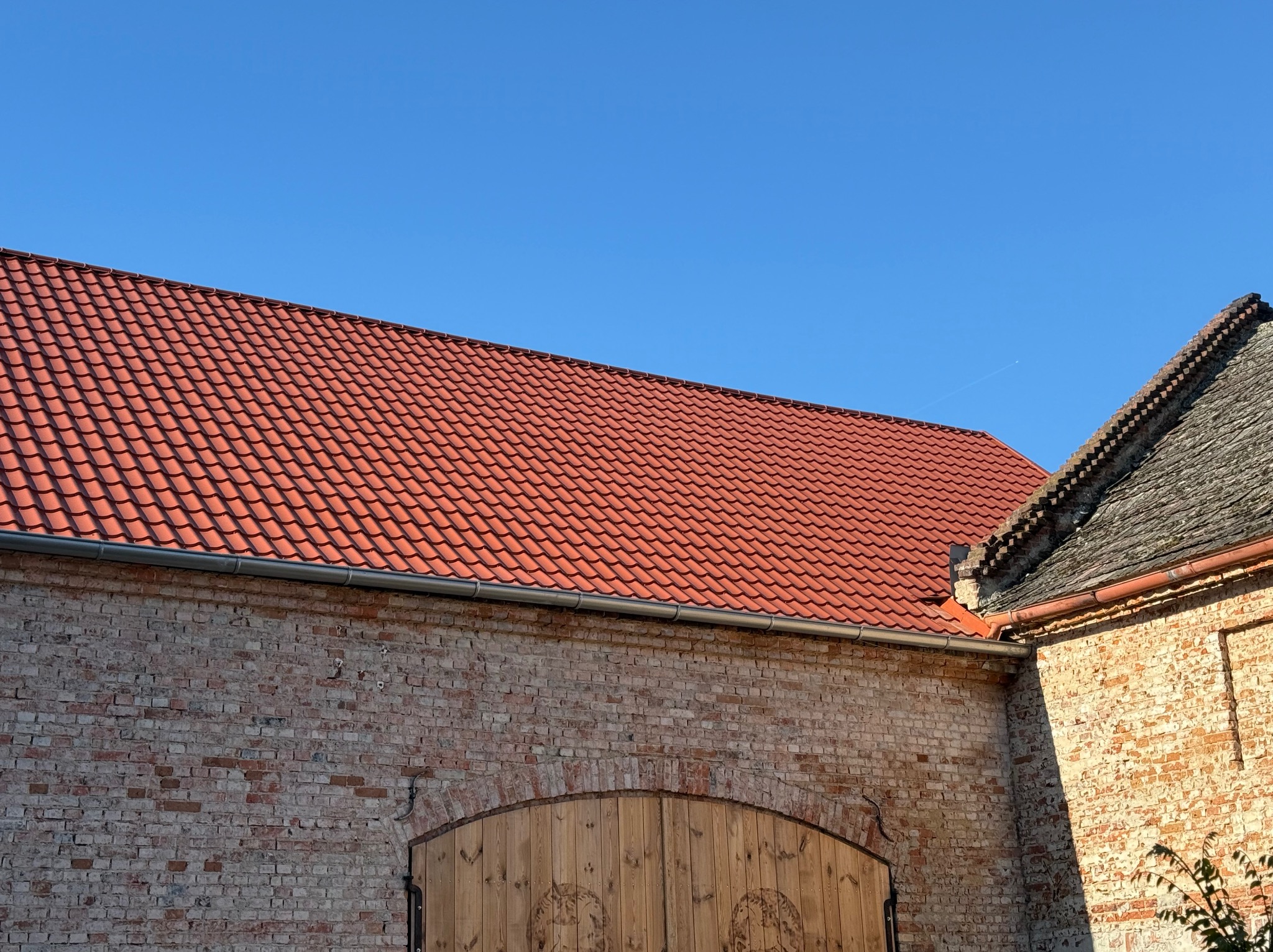 Czerwona dachówka na budynku z cegły, widoczne rynny i fragment innego dachu z szarej dachówki. Błękitne niebo w tle.