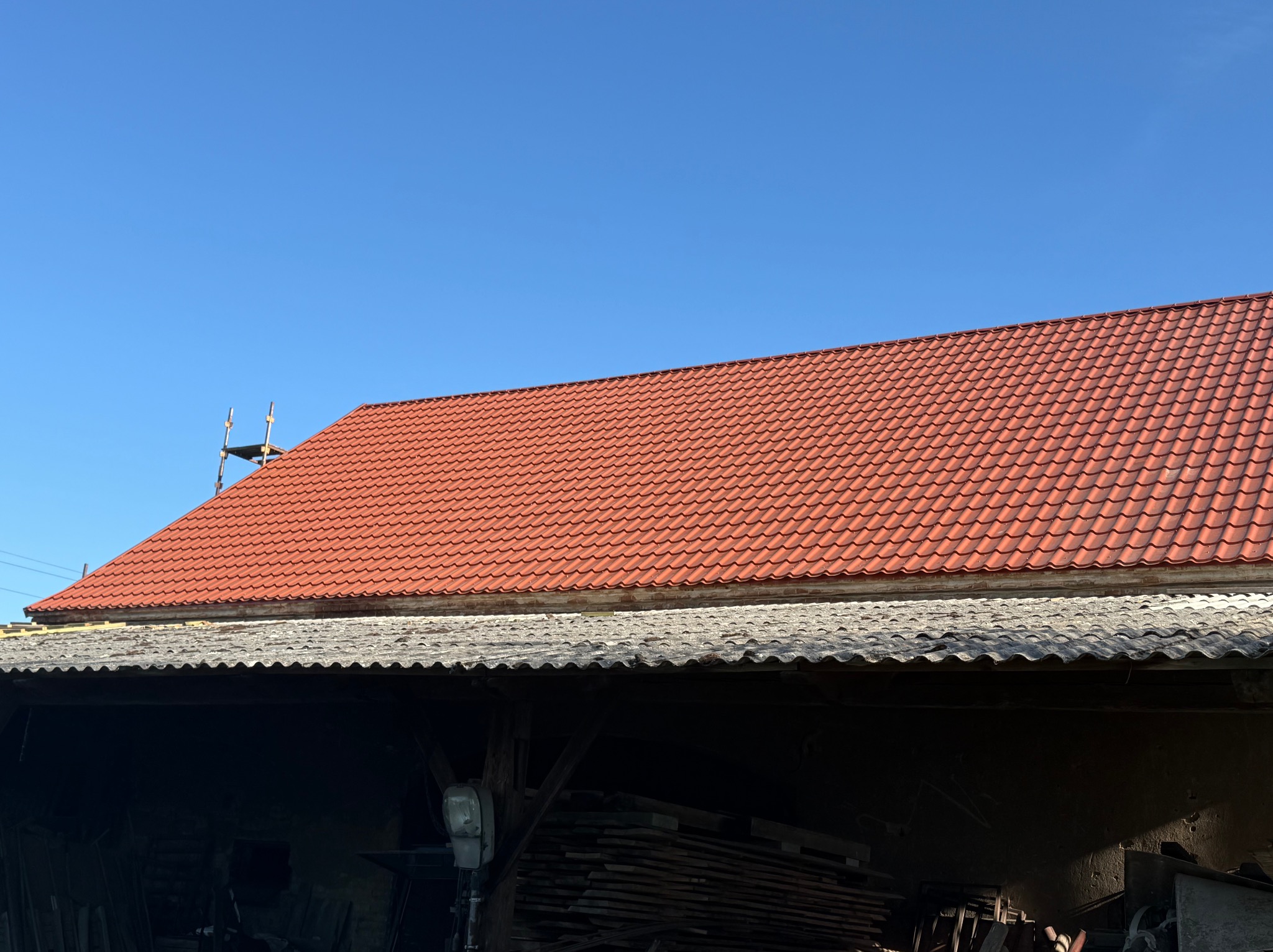 Czerwony dach z dachówki ceramicznej na budynku, widoczny fragment starego dachu i rusztowanie. Błękitne niebo w tle.