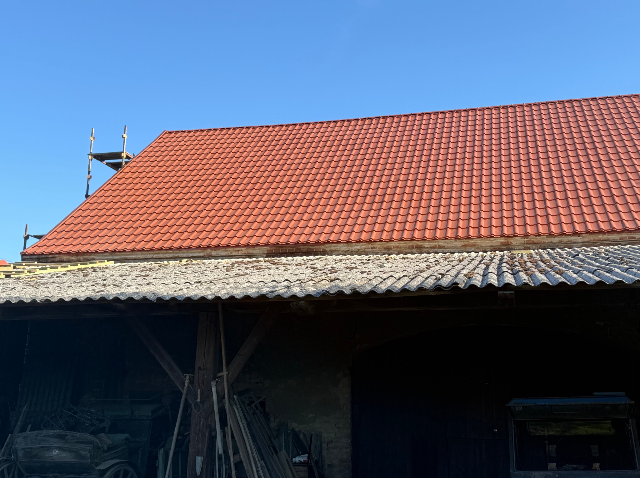 Czerwony dach z dachówki ceramicznej po wymianie, widoczny fragment starego dachu z eternitu oraz rusztowanie przy kominie na tle błękitnego nieba.