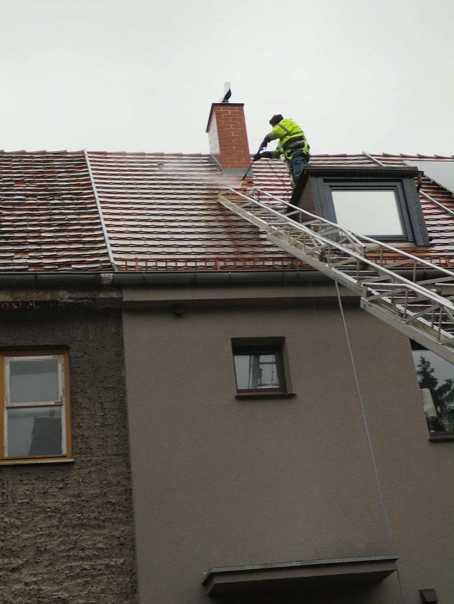 Pracownik w jaskrawej kurtce czyści dach z drabiny za pomocą myjki ciśnieniowej. Na kominie siedzi figurka bociana. Widoczne okno dachowe i fragment elewacji budynku.
