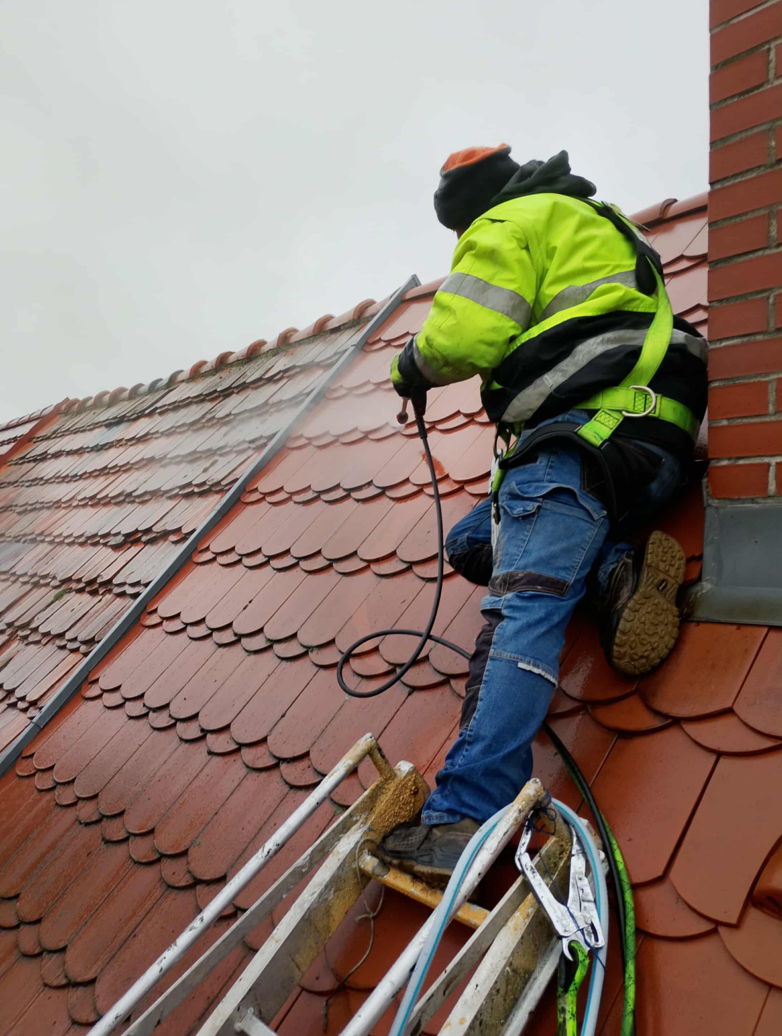 Pracownik w uprzęży, z drabiny, myje dach z czerwonej dachówki za pomocą myjki ciśnieniowej. Widoczny fragment komina z czerwonej cegły.