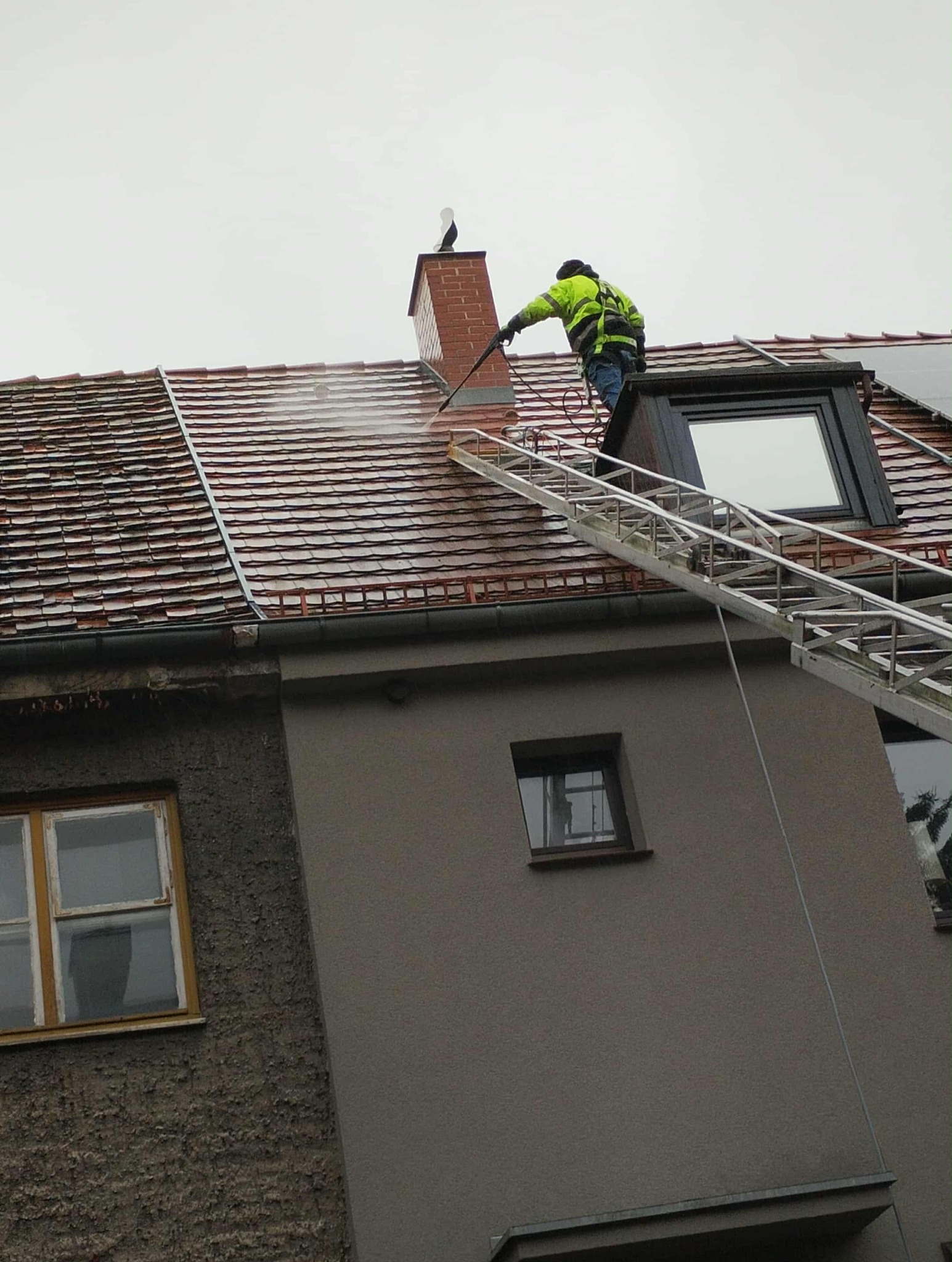 Pracownik w jaskrawej kurtce czyści dach z czerwonej dachówki za pomocą myjki ciśnieniowej, stojąc na drabinie. Na kominie siedzi figurka bociana.
