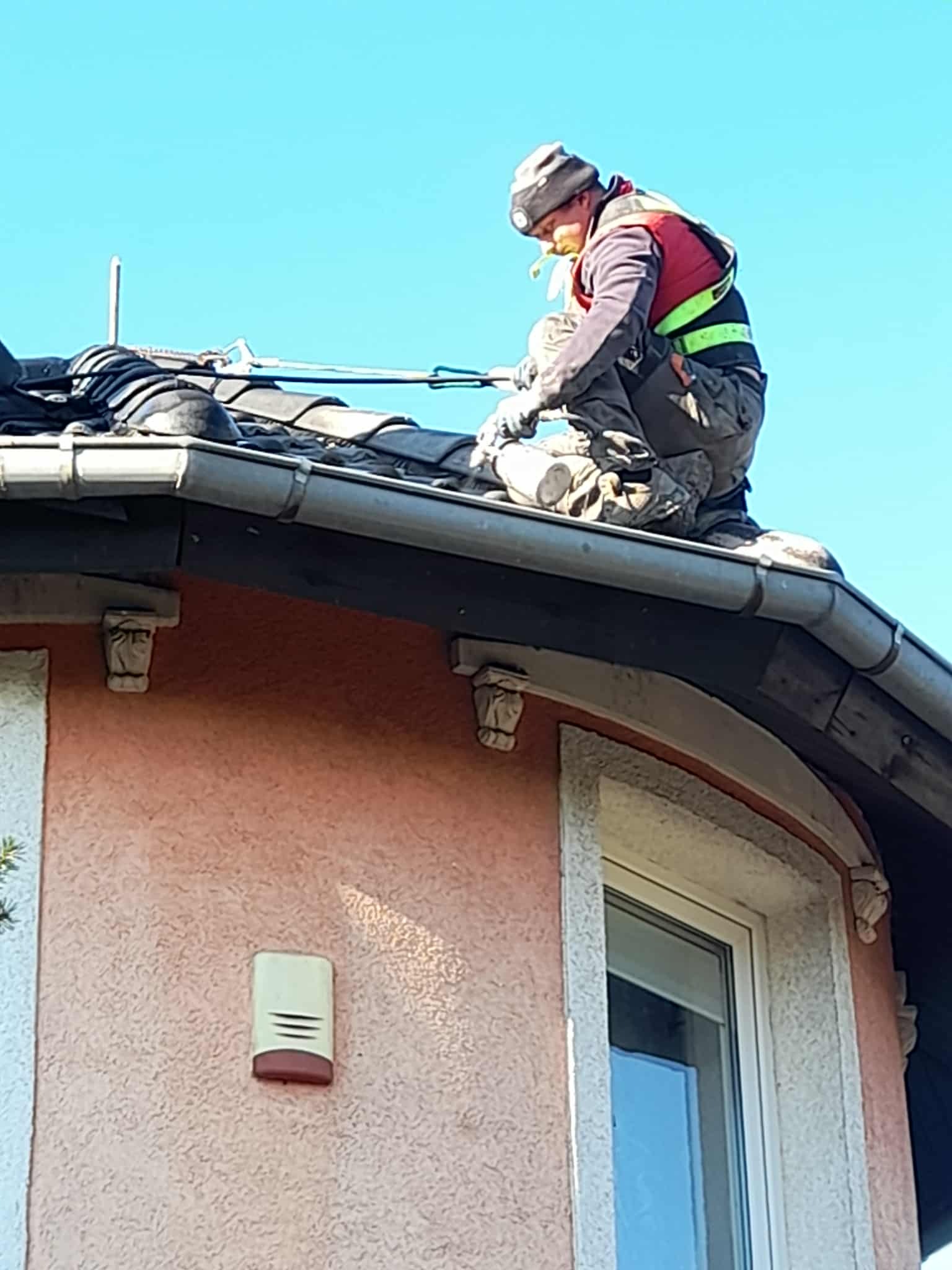 Pracownik w kamizelce na dachu z ciemnej dachówki, naprawia rynnę. Widoczny fragment elewacji budynku z oknem i ozdobnymi elementami.