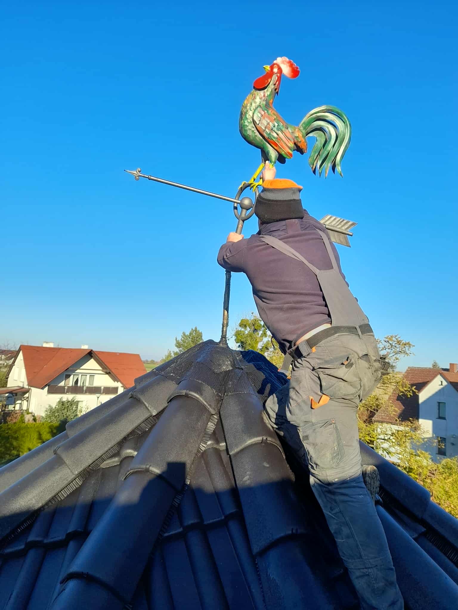 Dekarz montuje ozdobnego, kolorowego koguta-wiatrowskaz na szczycie ciemnego, ceramicznego dachu domu jednorodzinnego na tle błękitnego nieba.