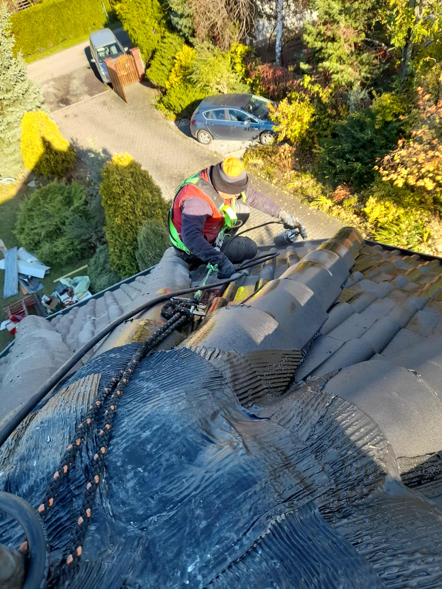 Pracownik w kasku i kamizelce, zabezpieczony linami, maluje dachówkę na ciemny kolor za pomocą pistoletu natryskowego. Widok z góry na częściowo pomalowany dach.