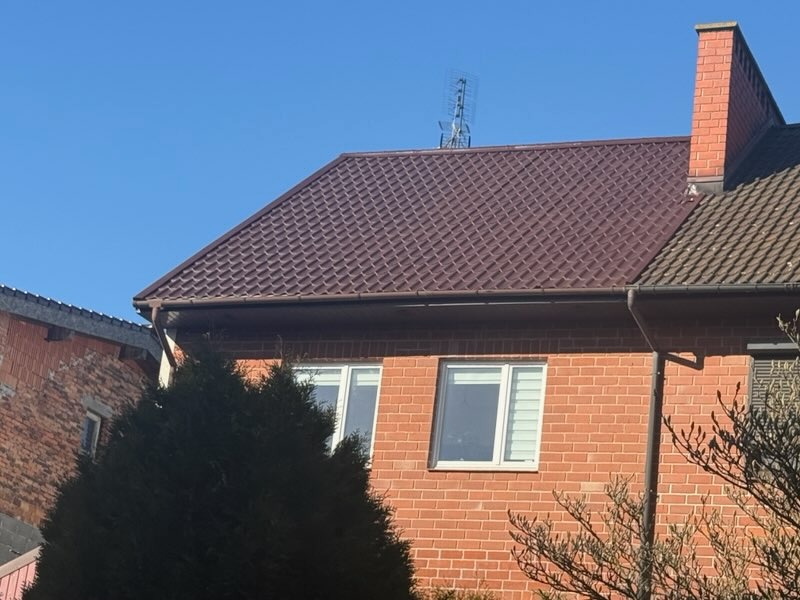 Ciemnobrązowy dach z blachy trapezowej na ceglanym domu z kominem i anteną, widok z dołu w słoneczny dzień.