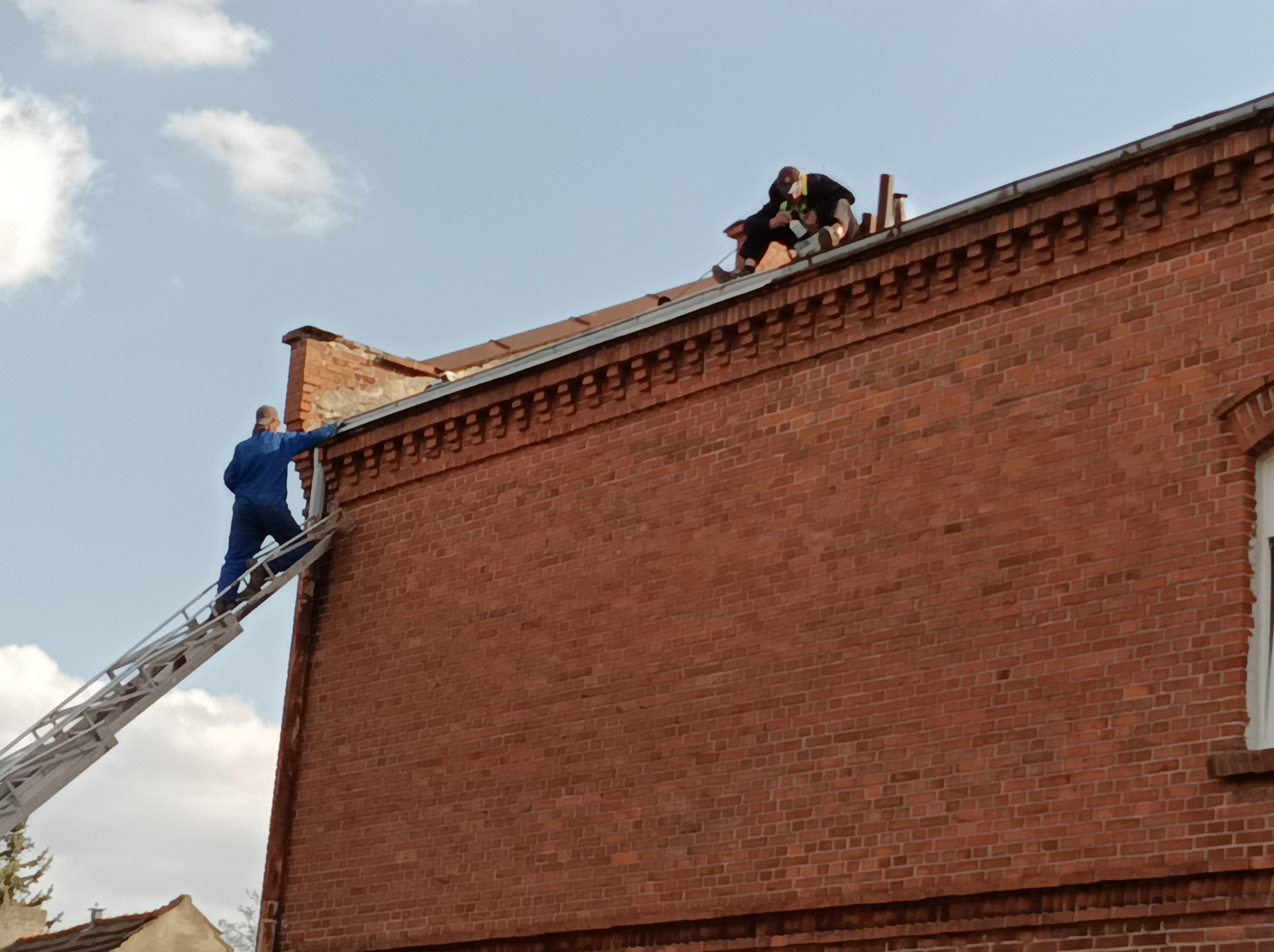 Dwóch dekarzy na dachu ceglanego budynku podczas pracy. Jeden wchodzi po drabinie, drugi pracuje na dachu. Błękitne niebo z chmurami w tle.