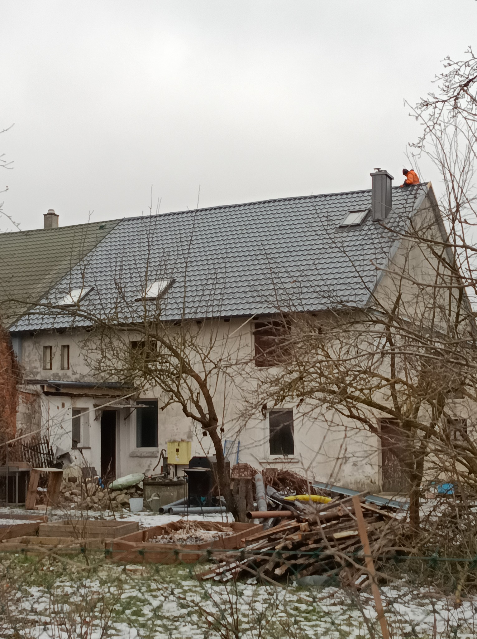 Pracownik w pomarańczowej kurtce na szarym dachu domu z lukarnami. W tle pochmurne niebo, a na pierwszym planie gałęzie drzew i resztki śniegu.
