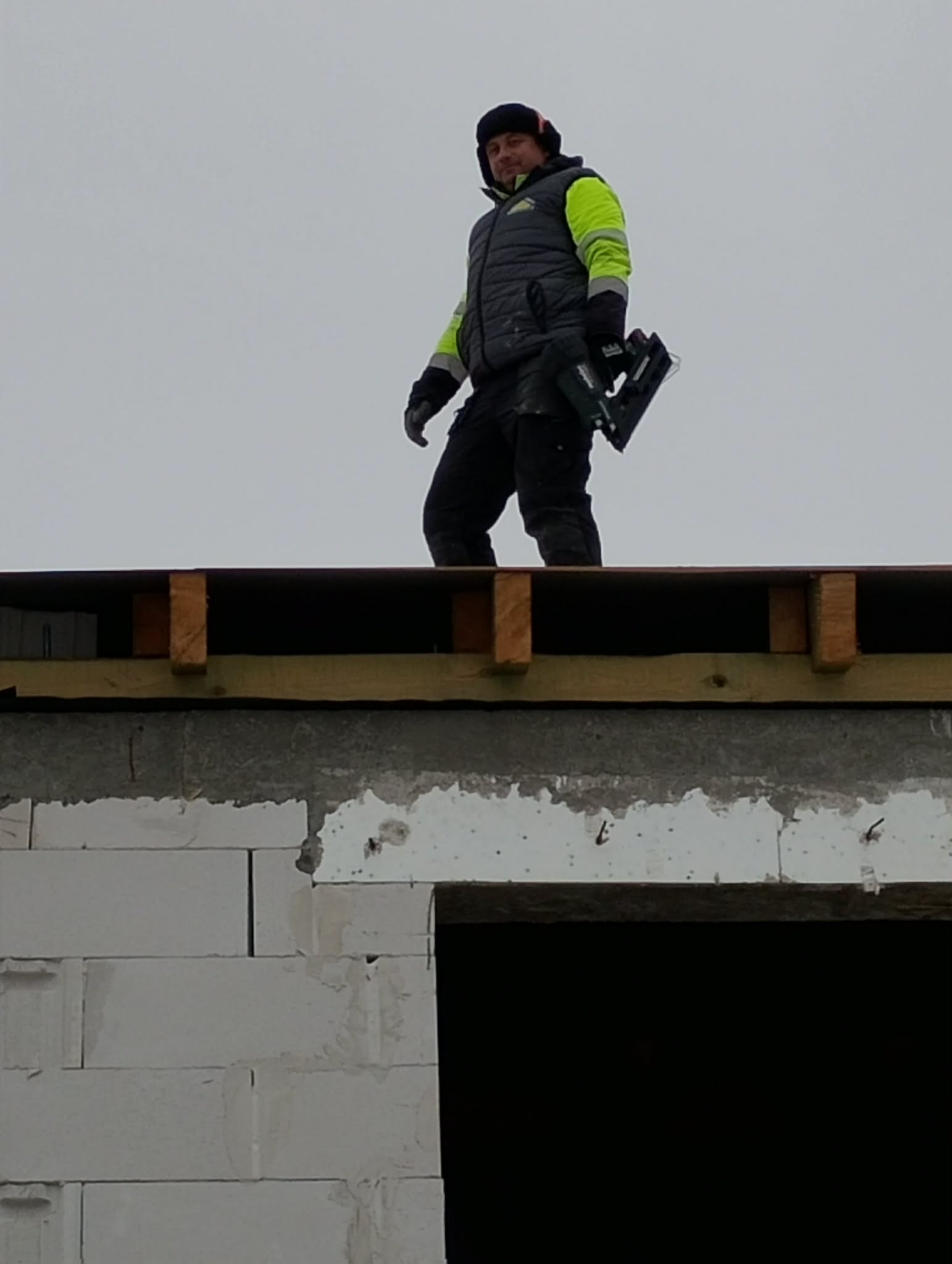 Dekarz z narzędziem na dachu w trakcie budowy, widok z dołu. Mężczyzna w odzieży roboczej, na tle szarego nieba. Widoczne bloczki betonowe i drewniana konstrukcja dachu.